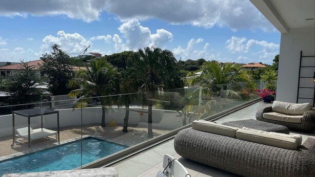 A modern outdoor living area with a pool, wicker furniture, and a glass railing overlooking a residential neighborhood. - Luxury 4 bed Villa - Private Pool - Sleeps 8 (Jan Thiel)