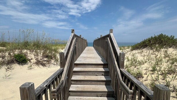 Une promenade en bois menant à une plage bordée de dunes de sable et d’une végétation clairsemée.