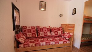 Un divano di legno con una fodera a motivi rossi e bianchi, un armadietto di legno e un letto a castello in una stanza dalle pareti chiare.