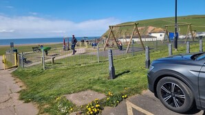 Un parque con juegos infantiles, un coche y vista al mar.
