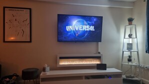 Una sala de estar moderna con un televisor de pantalla plana mostrando 'UNIVERSAL', un mueble blanco de entretenimiento debajo, una pequeña mesa con una planta, y una escalera con una planta en la parte superior.