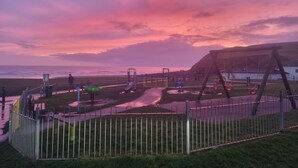 Un parque infantil con columpios y toboganes, un área cercada y vistas a la playa y al océano al atardecer.