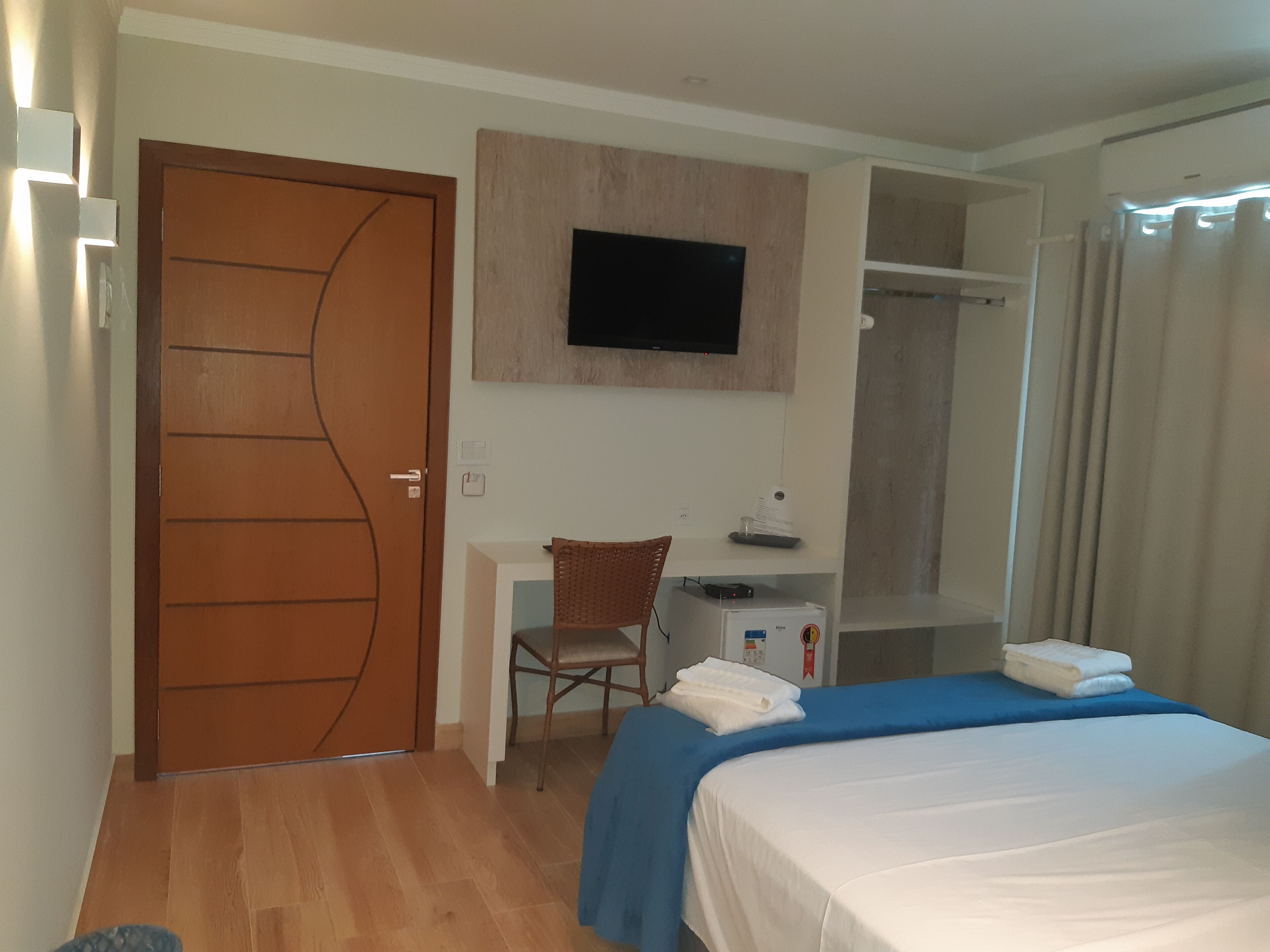 Quarto de hotel com porta de madeira, uma escrivaninha com cadeira, cama com colcha azul, televisão presa à parede e área de chuveiro.