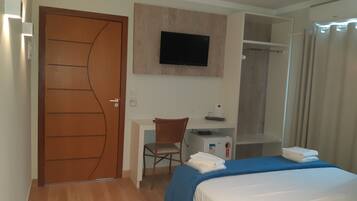 Quarto de hotel com porta de madeira, uma escrivaninha com cadeira, cama com colcha azul, televisão presa à parede e área de chuveiro.