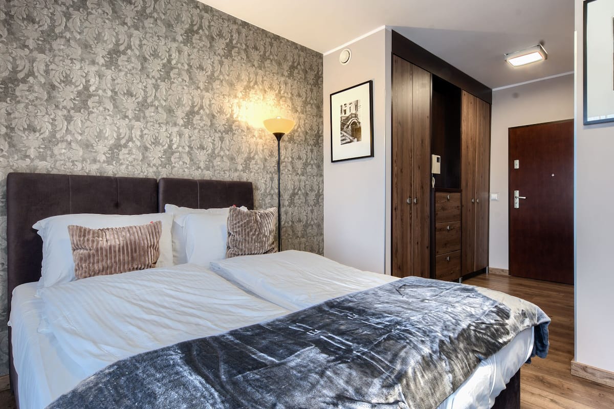 A bedroom with a large bed, a headboard, a wall-mounted lamp, a framed picture, and a wardrobe.