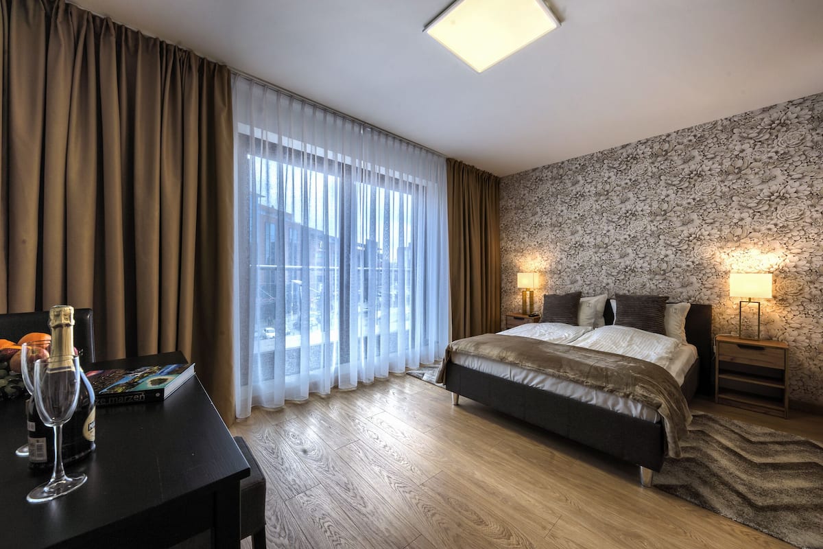 A hotel room with a bed, bedside lamps, a wooden floor, a window with sheer curtains, and a wall with a floral pattern.