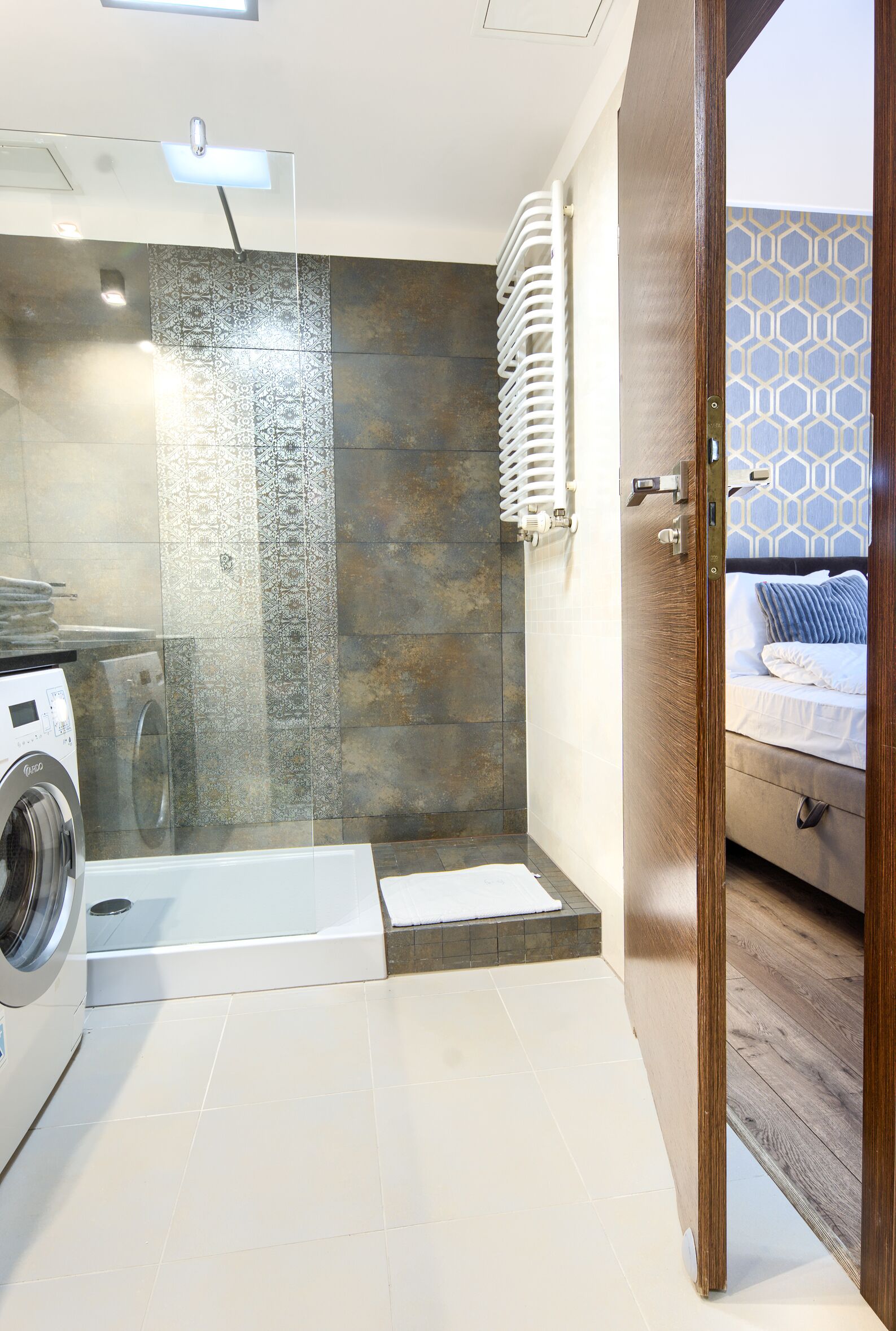 A modern bathroom with a glass shower enclosure, a built-in washing machine, and a wooden door leading to a bedroom with patterned wallpaper.