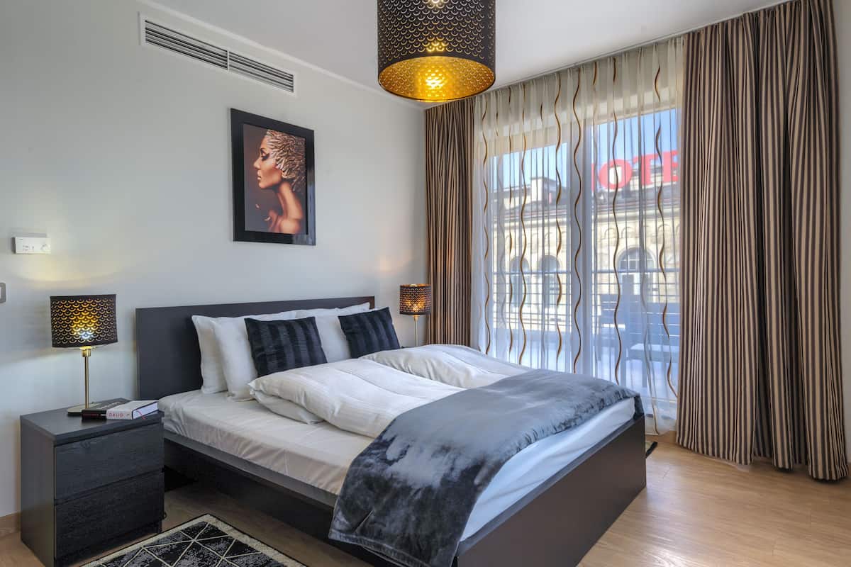 A modern bedroom with a large bed, bedside lamps, a framed artwork, and a view through curtains.