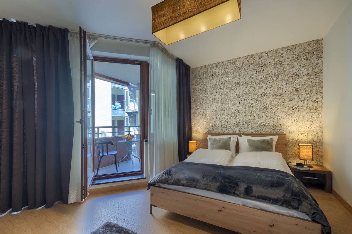 A bedroom with a wooden bed, a nightstand with a lamp, a balcony with a table and chairs, and a patterned wall.