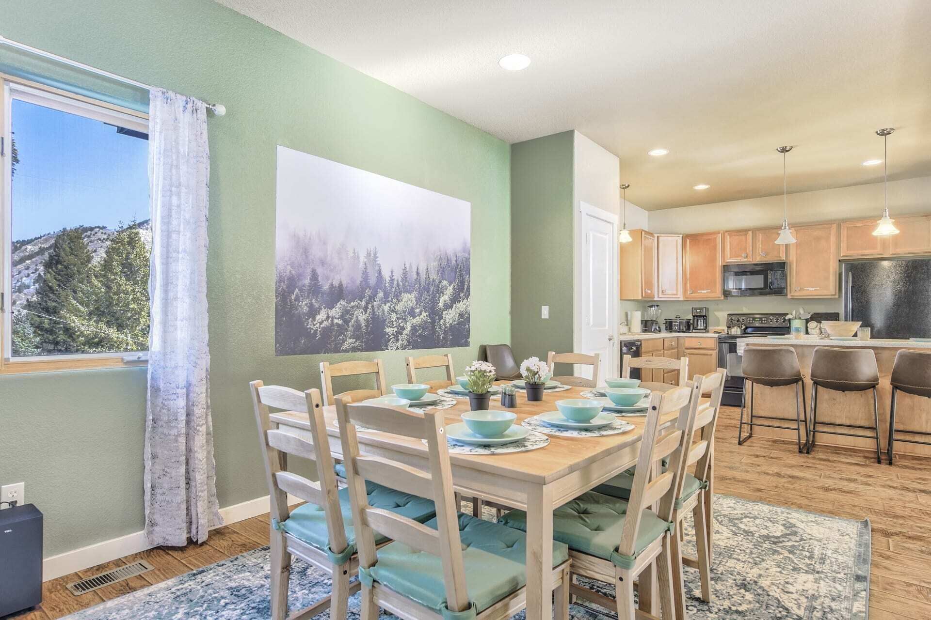 A dining area with a table set for four, wooden chairs with green cushions, a large window with curtains, and a kitchen with wooden cabinets and stainless steel appliances.