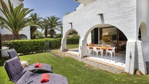 Un patio con due poltrone e un tavolo, un edificio bianco con archi e palme sullo sfondo.