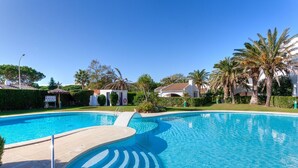 Piscina azzurra e cristallina con trampolino, circondata da palme e un edificio bianco con tetto rosso.