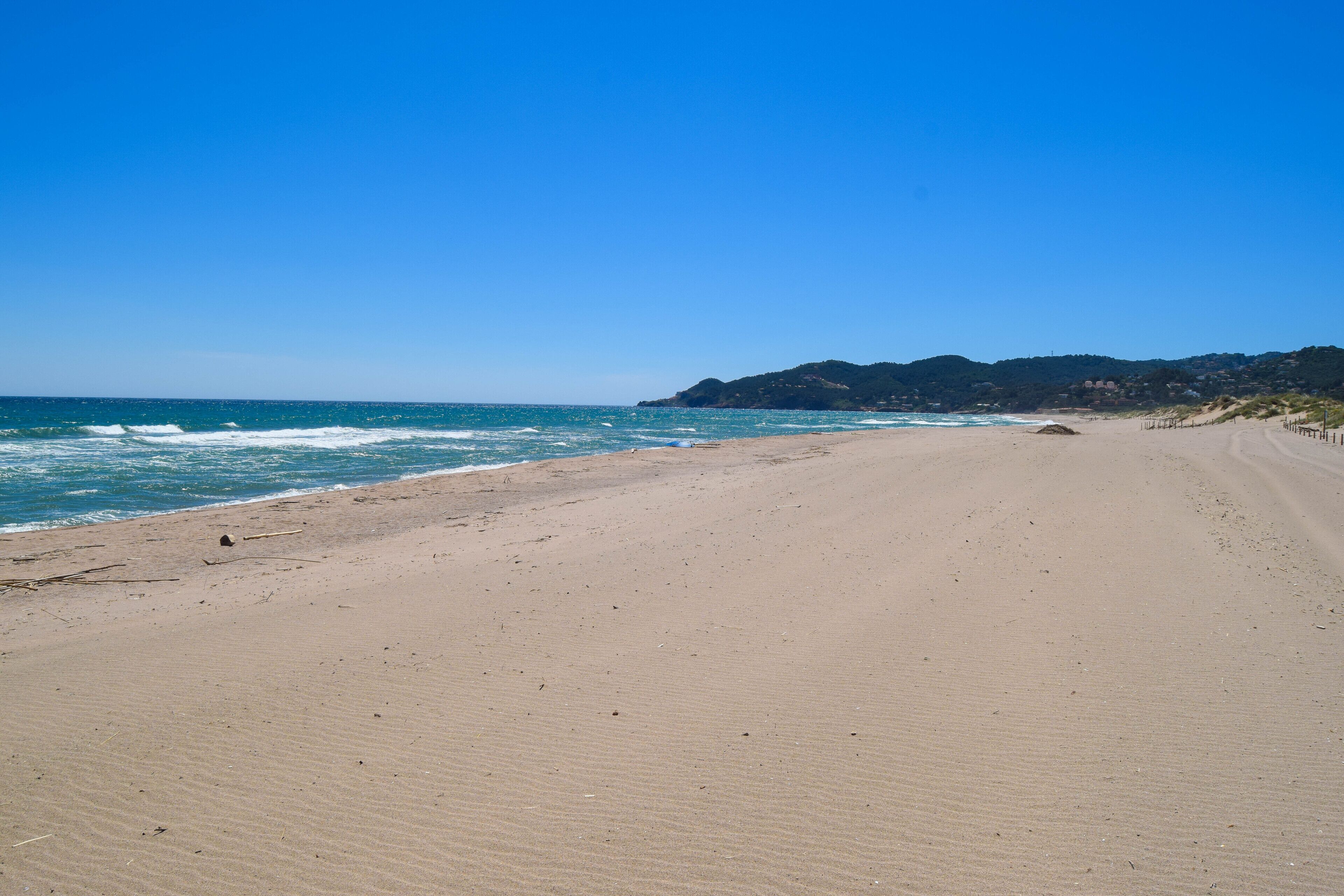 A wide sandy beach with gentle waves, a clear blue sky, and a distant hill with buildings.