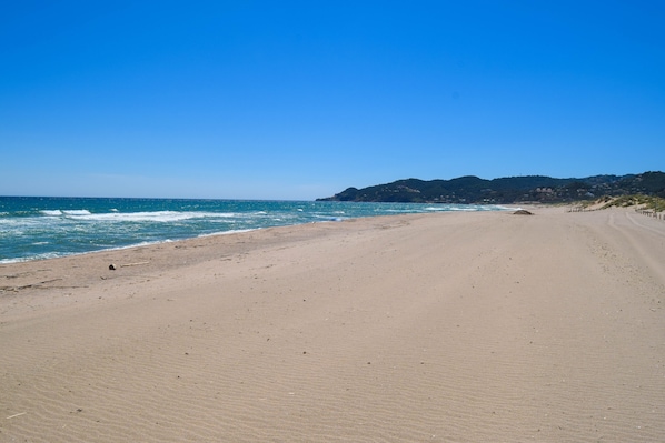 A wide sandy beach with gentle waves, a clear blue sky, and a distant hill with buildings.