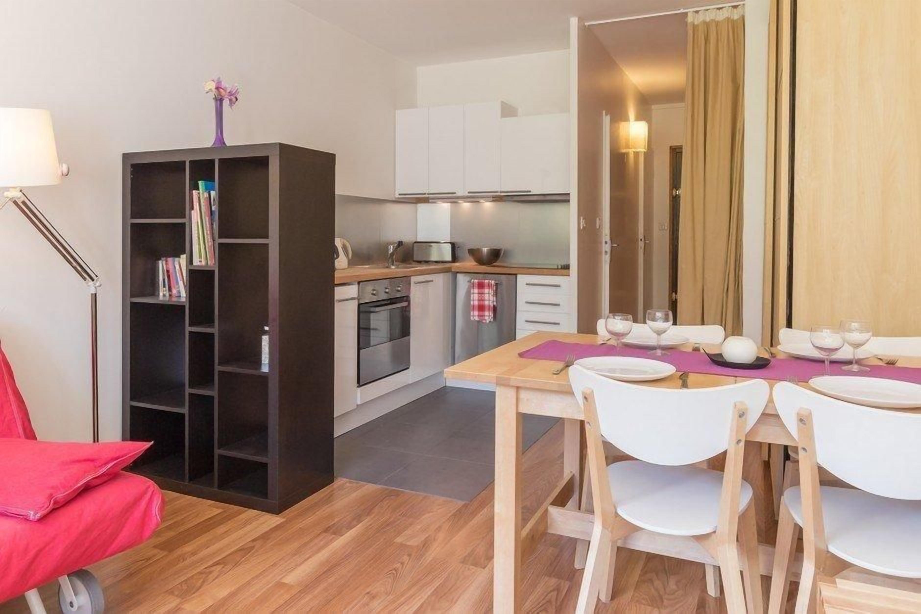 Una cocina compacta con armarios blancos, un comedor con una mesa dispuesta para dos personas y una estantería llena de libros.