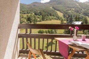 A balcony with a wooden railing, a table with a pink tablecloth, a chair, and a vase with pink flowers.