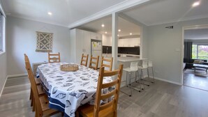 A dining area with a table set for six, wooden chairs, a woven basket, and a kitchen in the background. - Pirralea (Nelson Bay)