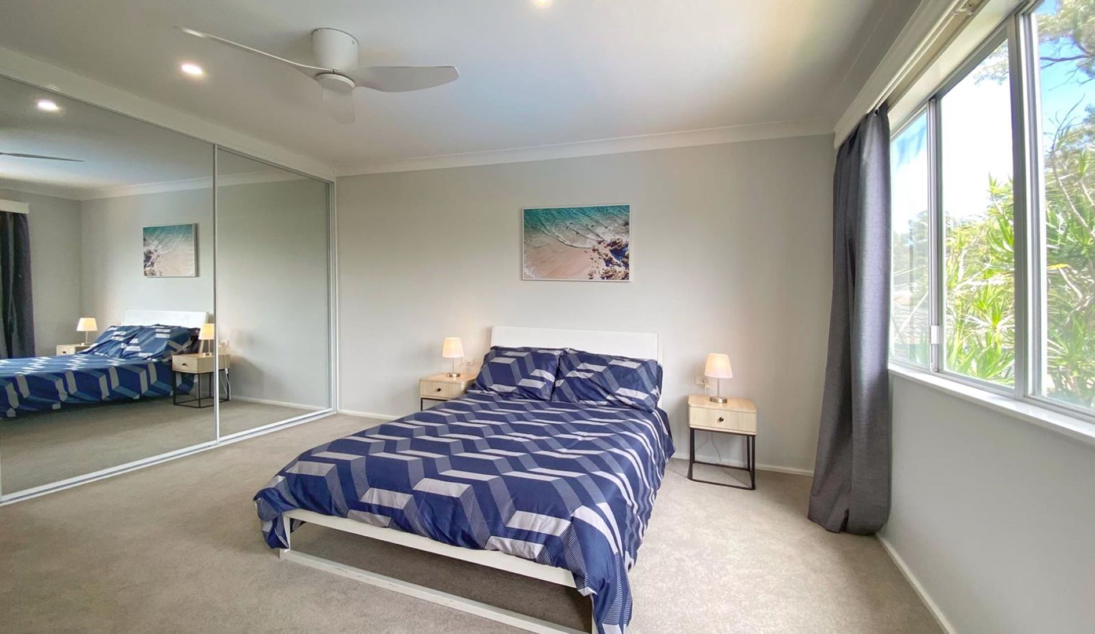 Ein Schlafzimmer mit einem Bett, zwei Nachttischen, einem Deckenventilator, einem großen Fenster mit Vorhängen und einem Wandbild.