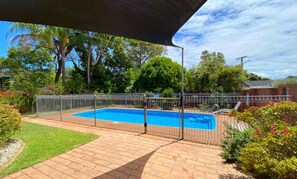 A rectangular swimming pool with a metal fence, surrounded by a brick patio and lush greenery.