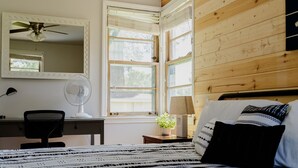 Une chambre à coucher comprenant un lit, un bureau avec une chaise, un ventilateur sur pied, un ventilateur de plafond et une fenêtre avec des stores.