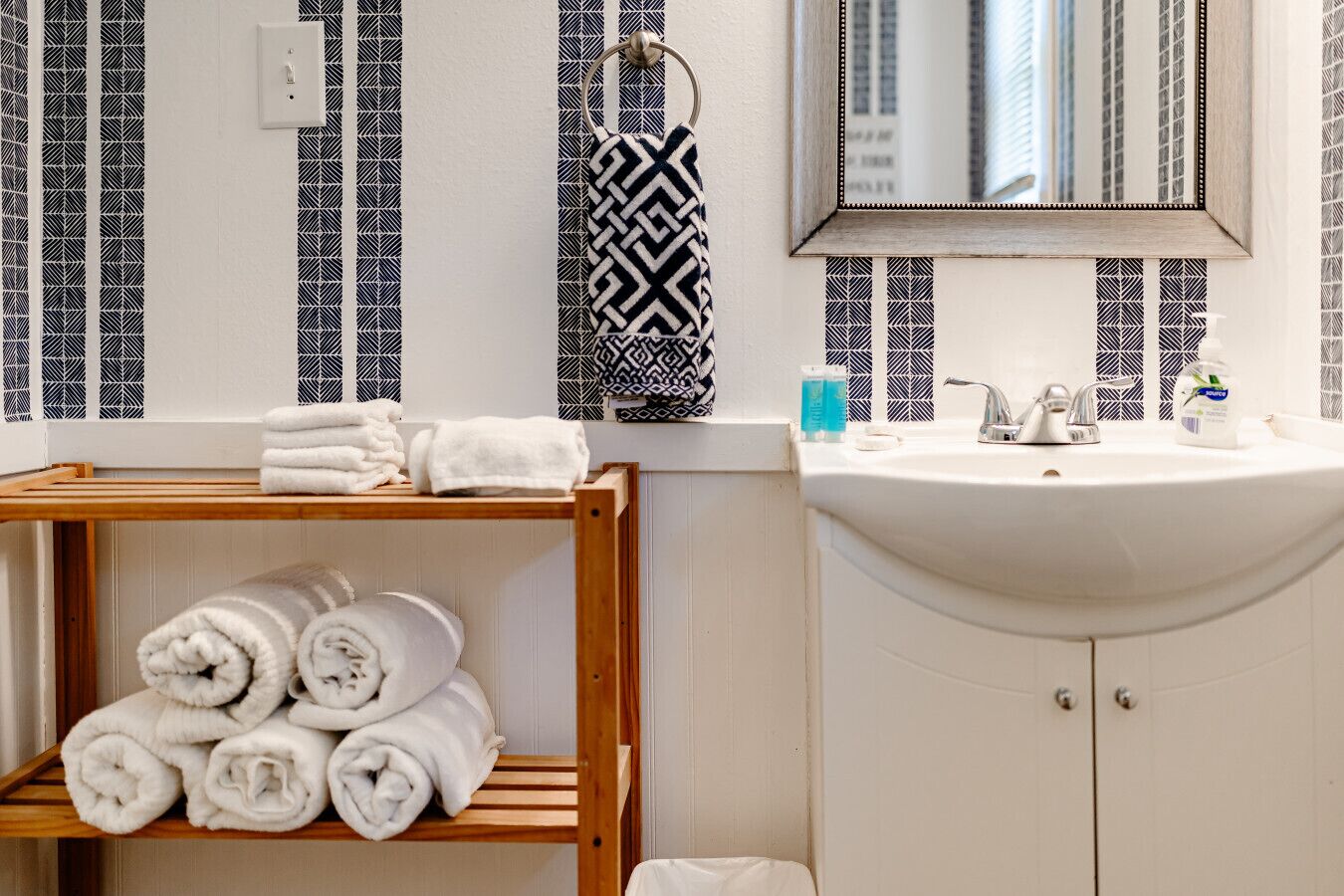 A bathroom with a wooden shelf holding rolled towels, a sink with toiletries, and a mirror above it.