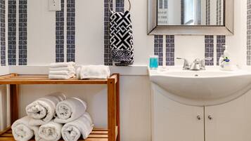 A bathroom with a wooden shelf holding rolled towels, a sink with toiletries, and a mirror above it.