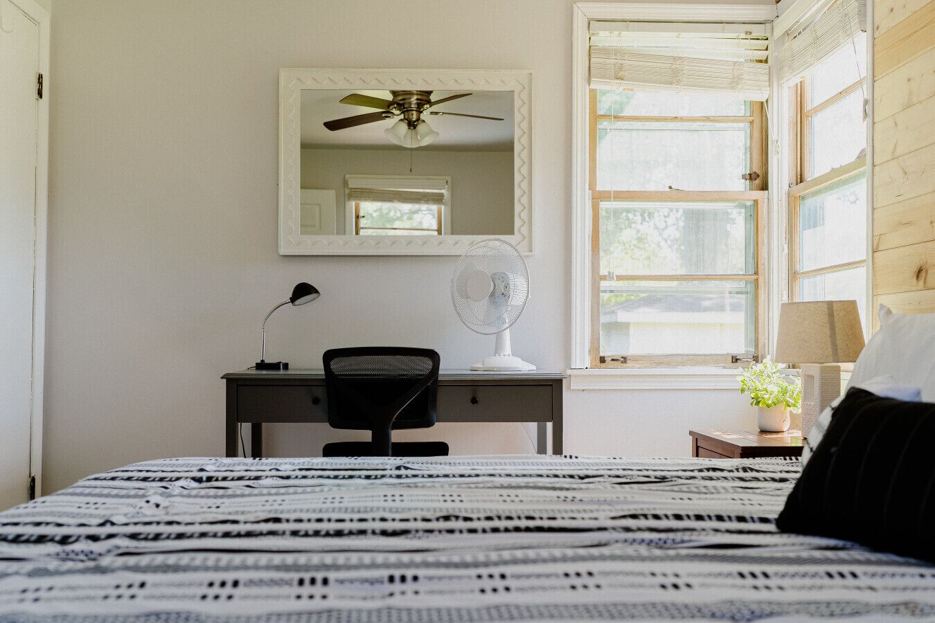 A bedroom with a bed, a desk, a chair, a ceiling fan, a mirror, and a window with blinds.