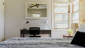 A bedroom with a bed, a desk, a chair, a ceiling fan, a mirror, and a window with blinds.