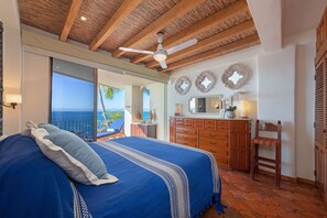 Quarto com cama grande, ventilador de teto, vigas de madeira, cômoda e vista para o oceano.