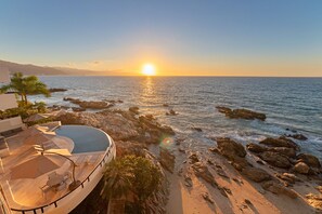 Vista do pôr do sol a partir de um terraço no telhado com piscina, com vista para o mar e a costa rochosa.