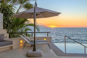 Uma piscina infinita com vista para o pôr do sol sobre o oceano, área de descanso com um grande guarda-sol e uma escadaria que leva à piscina.