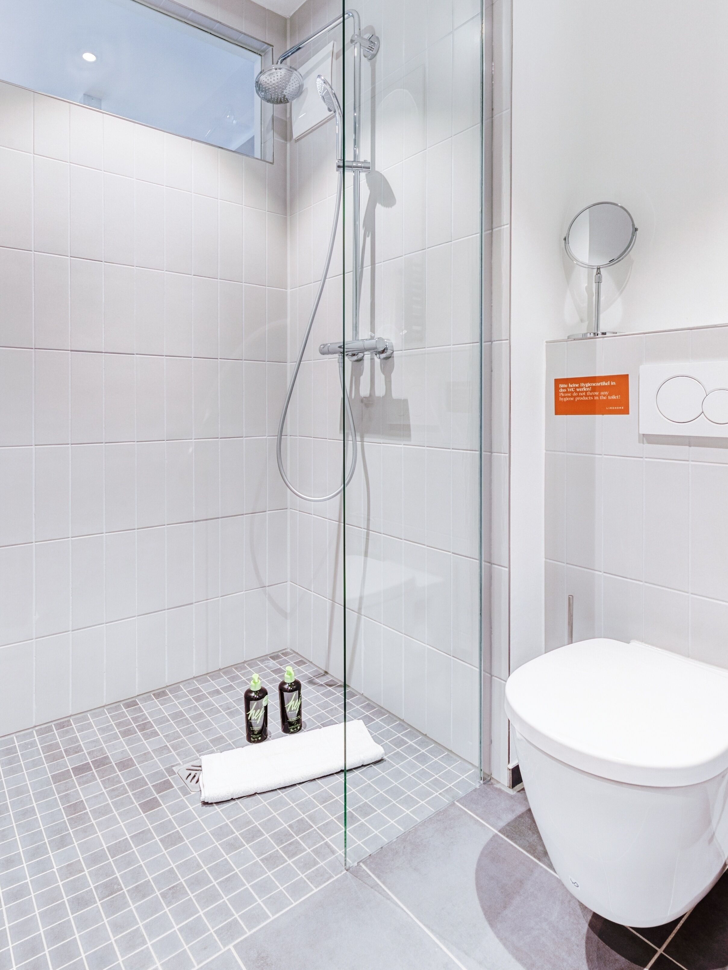 A modern bathroom with a glass-enclosed shower, a white toilet, and a small shelf with hygiene products.