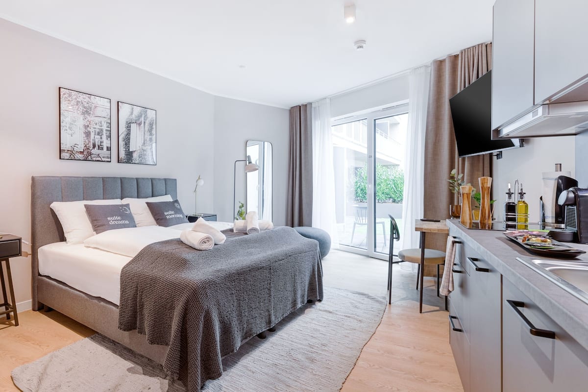 Quarto de hotel moderno com uma cama grande, televisão de tela plana, cozinha compacta e porta de vidro deslizante para o exterior.