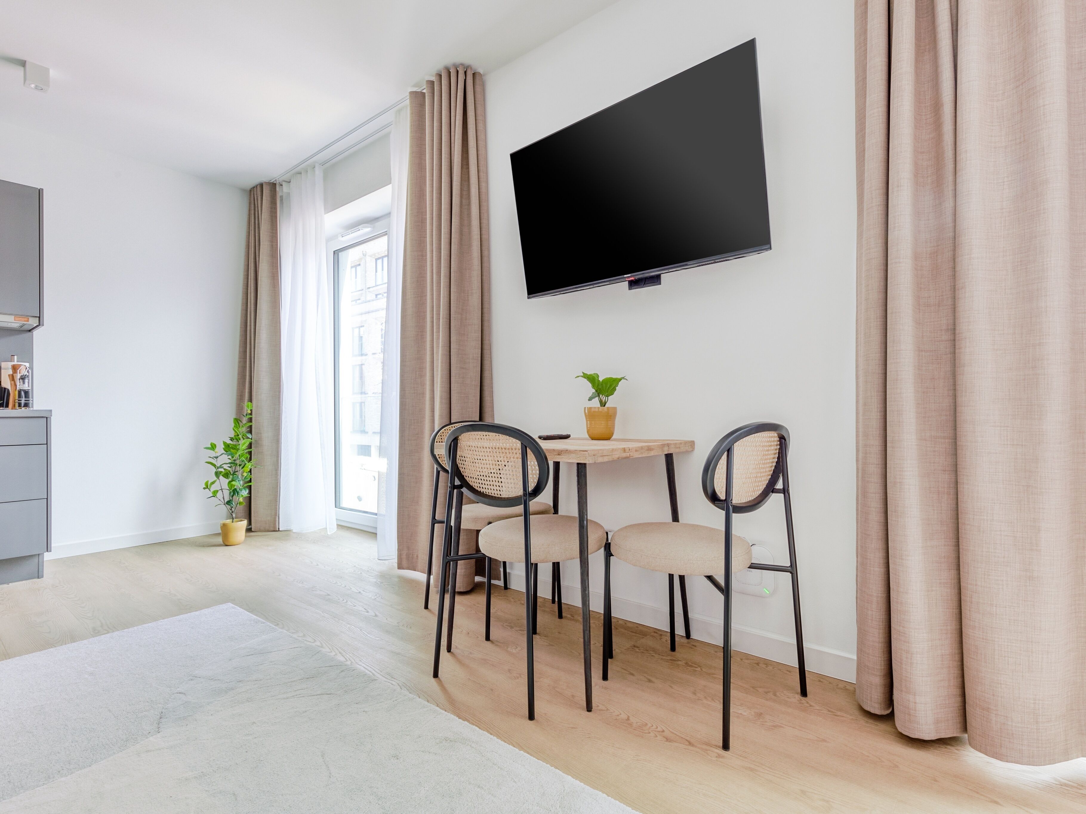 Un salon moderne avec un téléviseur à écran plat fixé au mur, une petite table ronde ornée d’un pot de fleurs et deux chaises à dossier tressé.