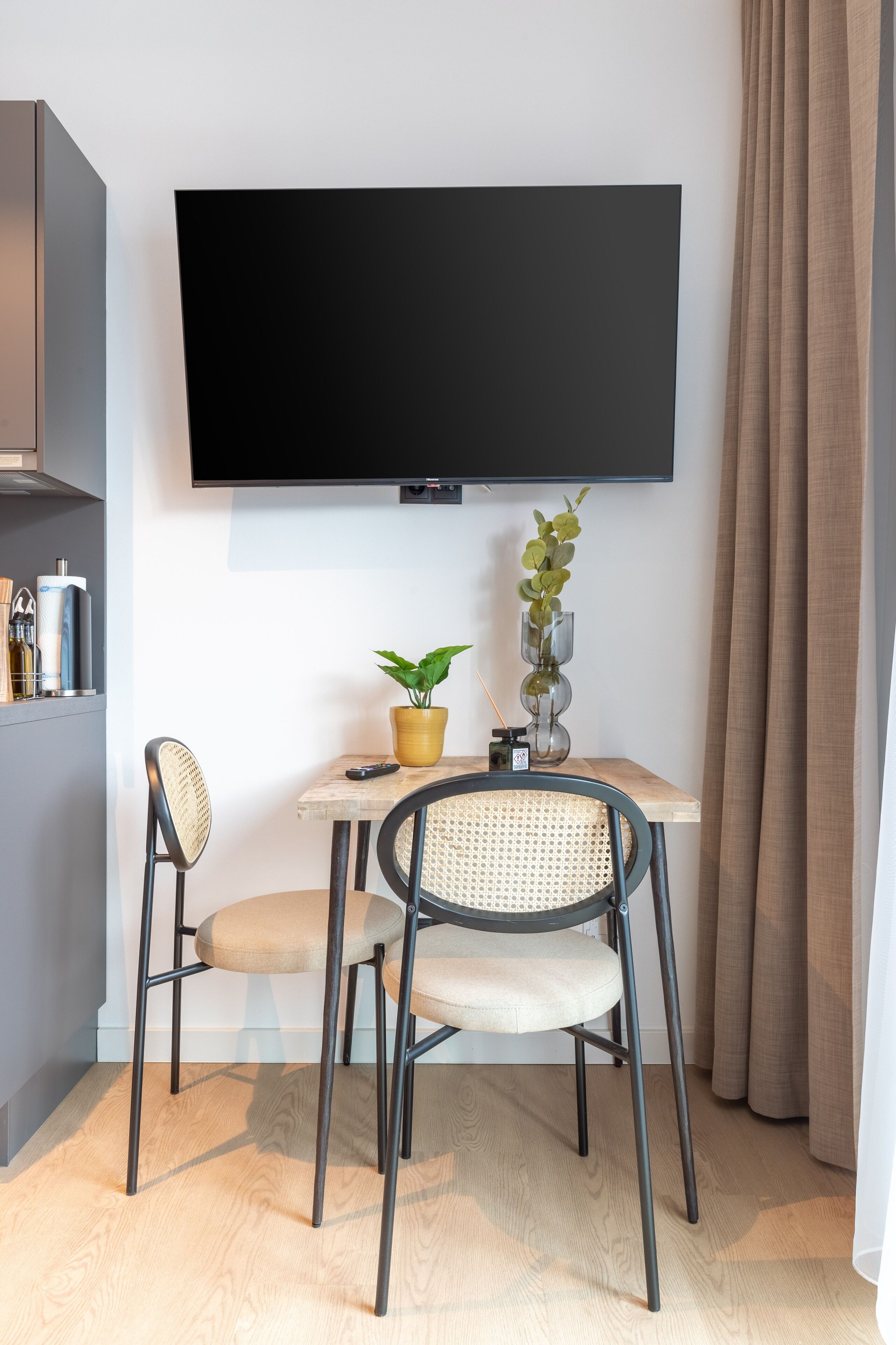 A modern living room with a flat-screen TV mounted on the wall, a small wooden table with two chairs, a potted plant, and a remote control.