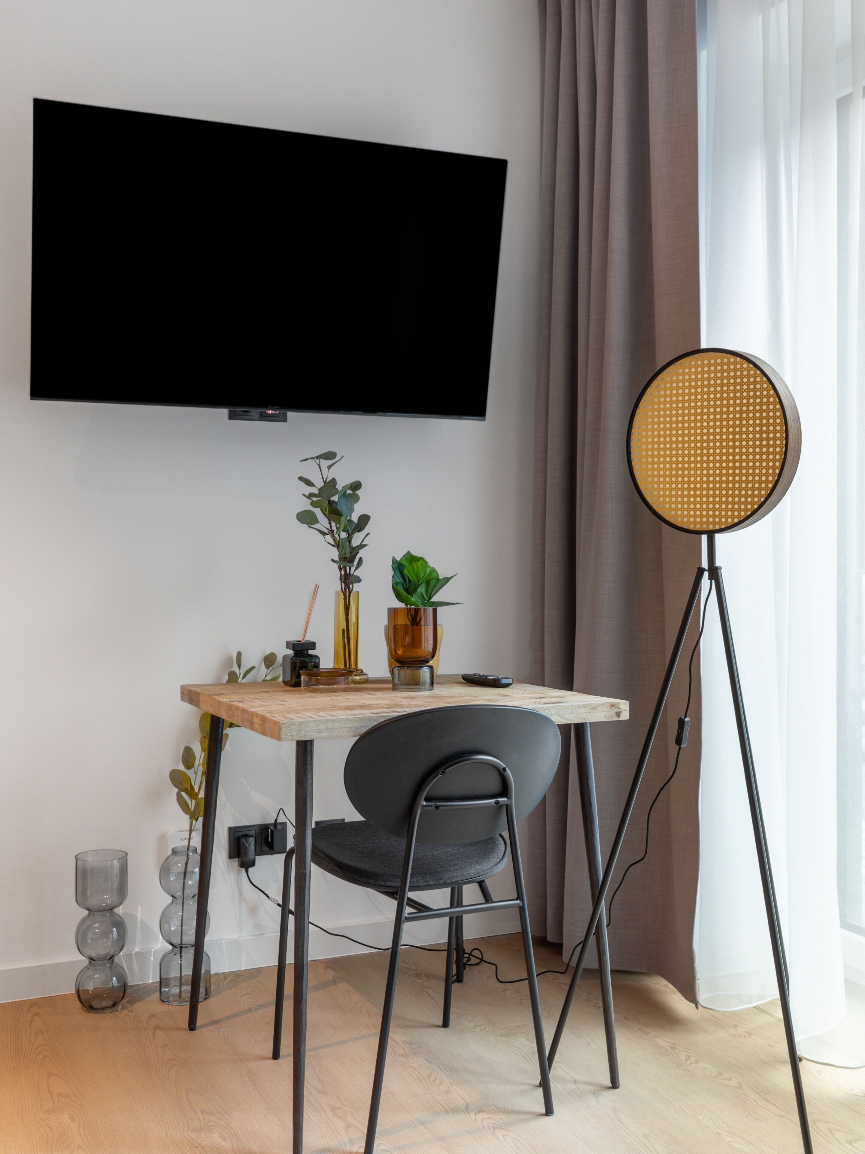 Una sala de estar moderna con un televisor de pantalla plana montado en la pared, una lámpara de pie redonda, un escritorio de madera con una silla y una planta en maceta.
