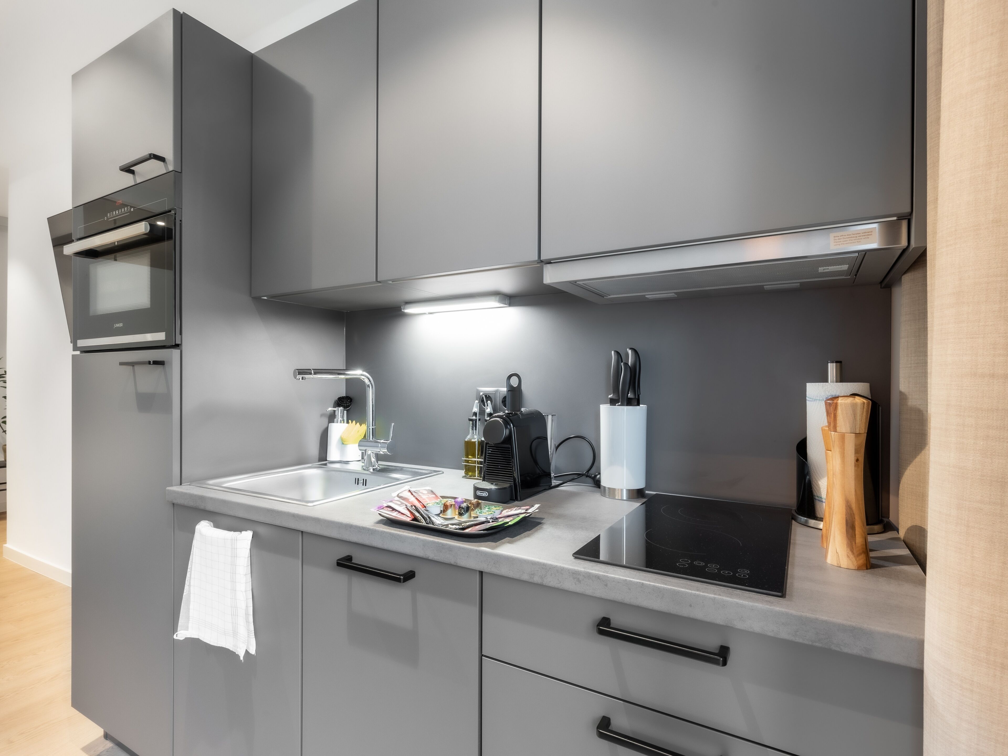 A modern kitchen with grey cabinets, a stainless steel sink, a black induction cooktop, and a built-in oven.