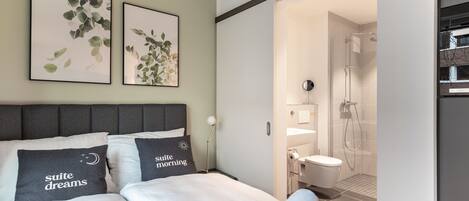 A modern bedroom with a bed, two pillows, a bedside lamp, and a bathroom visible through an open door.