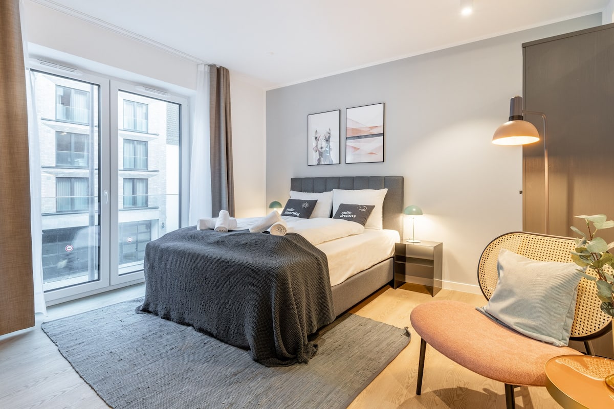 Quarto moderno com uma cama grande, mesa de cabeceira, uma cadeira e janela com cortinas.