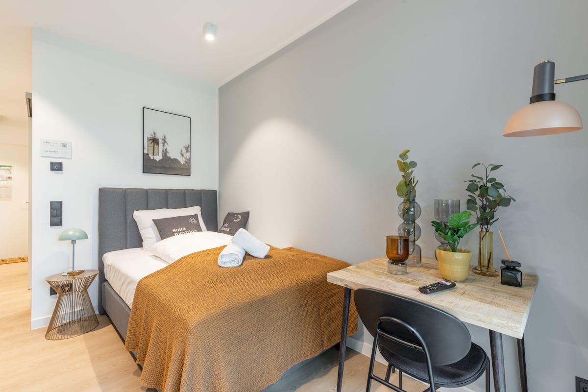 Quarto de hotel moderno com uma cama, uma escrivaninha, uma cadeira, uma luminária e um vaso de plantas.