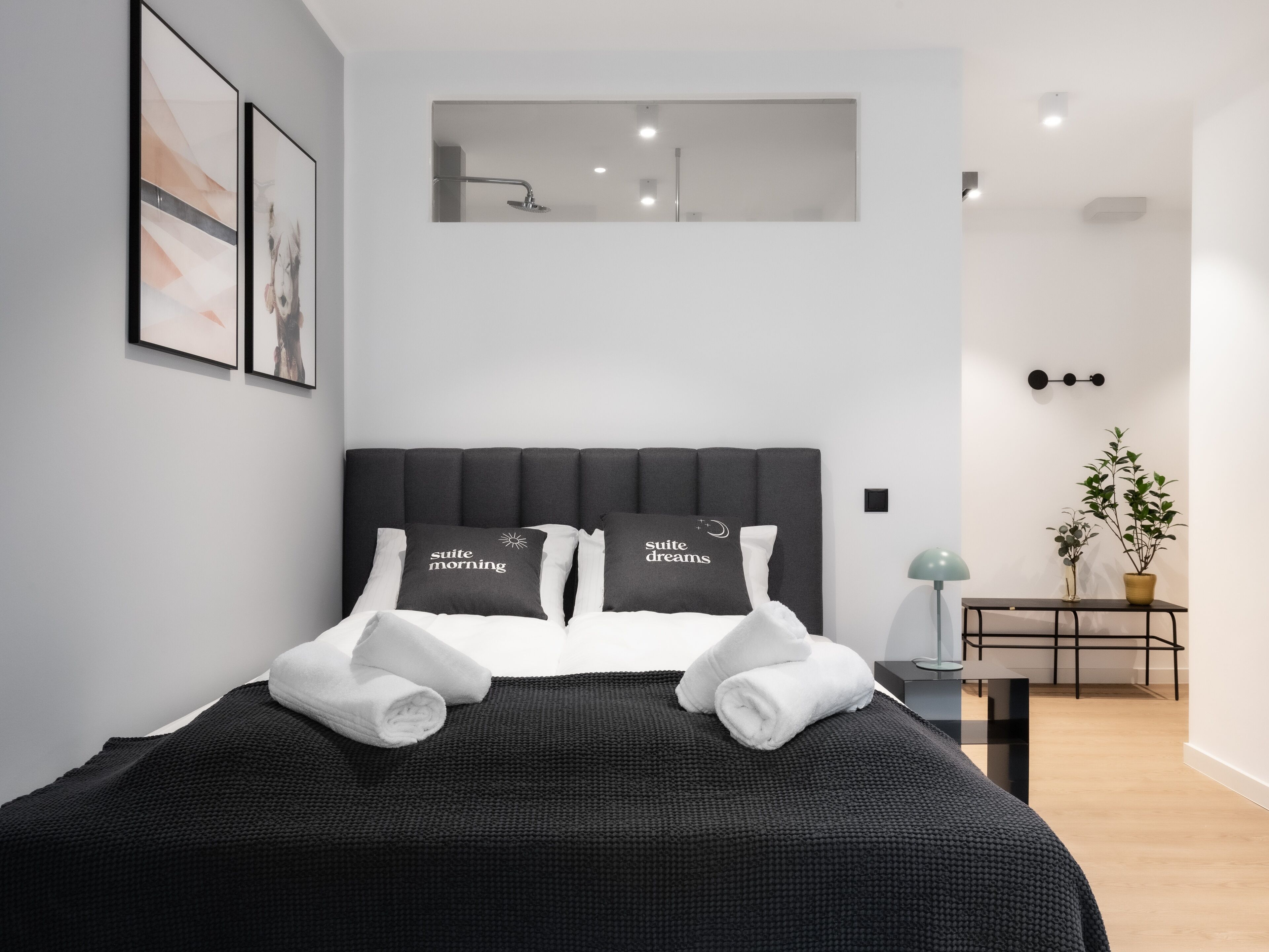 Quarto de hotel moderno com uma cama grande, duas almofadas com texto, uma pequena mesa com uma planta e um candeeiro fixado na parede.