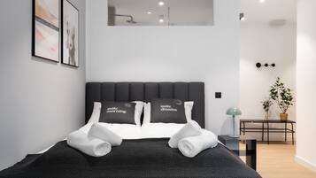 Quarto de hotel moderno com uma cama grande, duas almofadas com texto, uma pequena mesa com uma planta e um candeeiro fixado na parede.