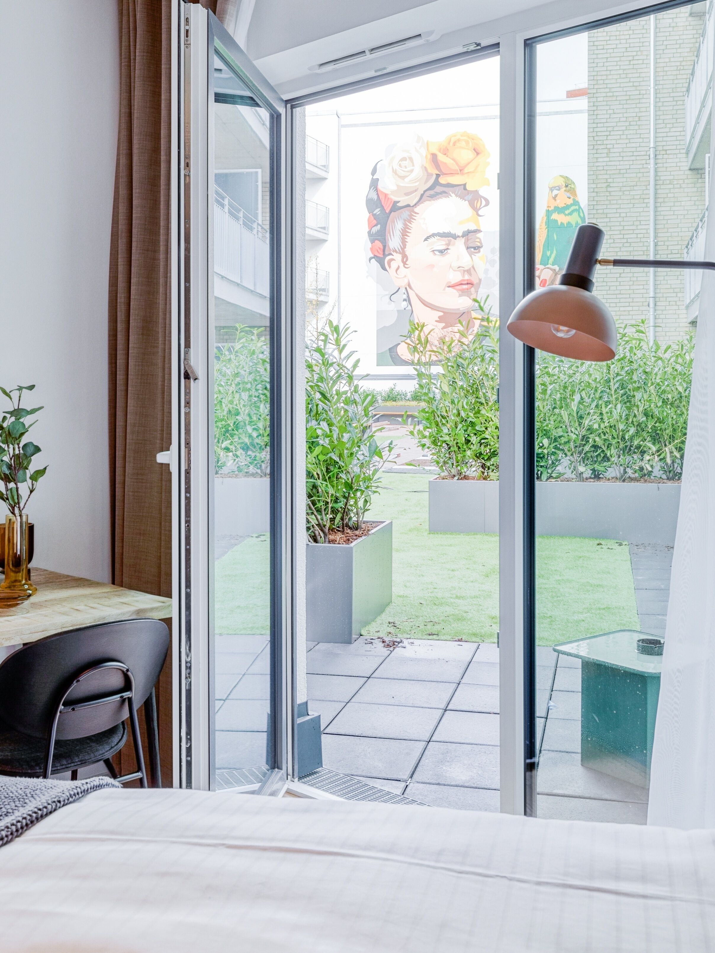 A modern hotel room with a large sliding glass door leading to a balcony with potted plants and a view of the cityscape.
