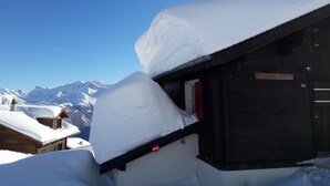 Un bâtiment enneigé, doté d’une façade en bois et d’une porte rouge, se dressant sur fond de montagnes couvertes de neige et d’un ciel bleu dégagé.