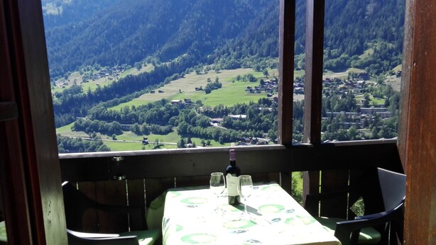 Une table recouverte d’une nappe, des chaises et une bouteille de vin, le tout sur un balcon donnant sur une vallée avec des montagnes en arrière-plan.