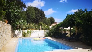 Ein rechteckiger Swimmingpool mit einer umlaufenden Holzterrasse, vor einer Baumlandschaft und blauem Himmel.