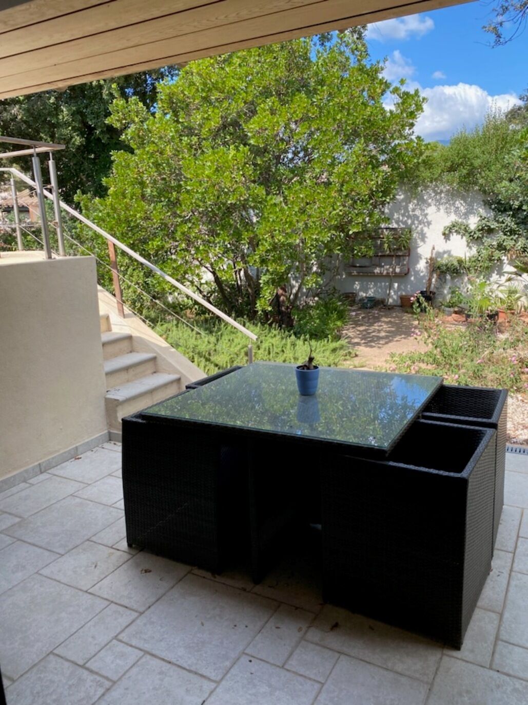 Un patio cubierto con una mesa rectangular y sillas, rodeado de vegetación y una escalera que conduce a un nivel superior.