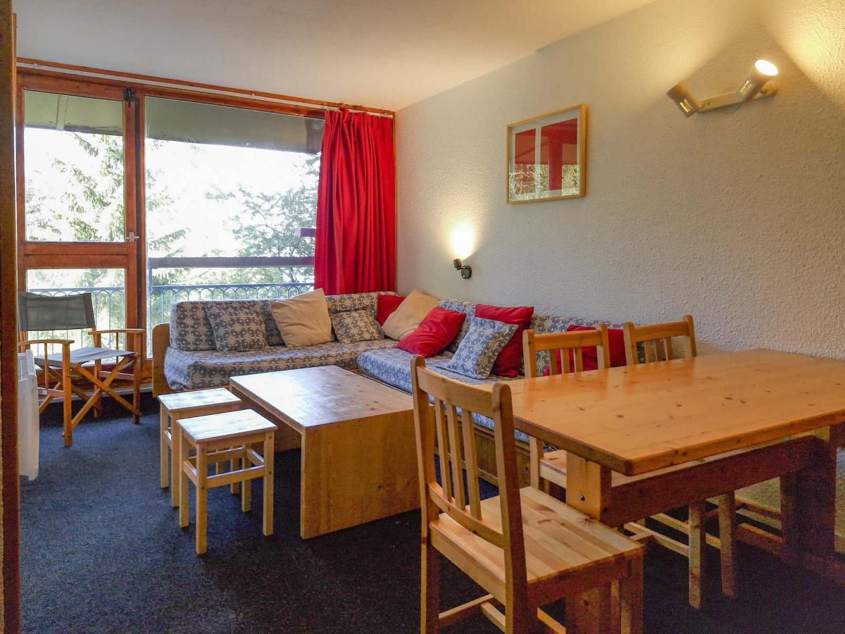 Et hyggeligt værelse med et træbord, stole, en sofa med mønstrede puder og et vindue med røde gardiner.
