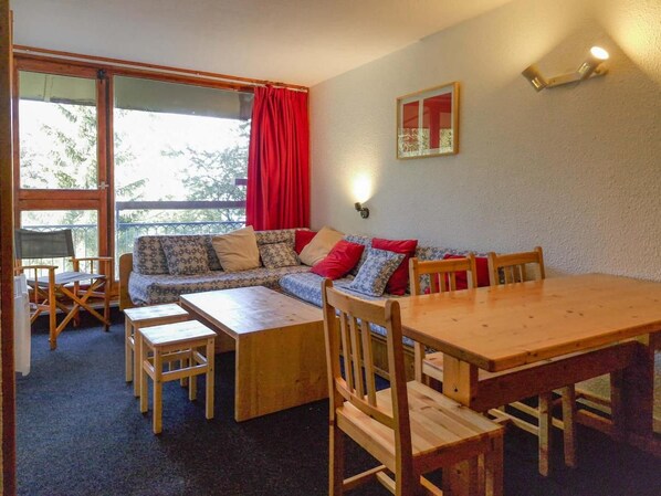 A cozy room with a wooden dining table, chairs, a sofa with patterned cushions, and a window with red curtains.