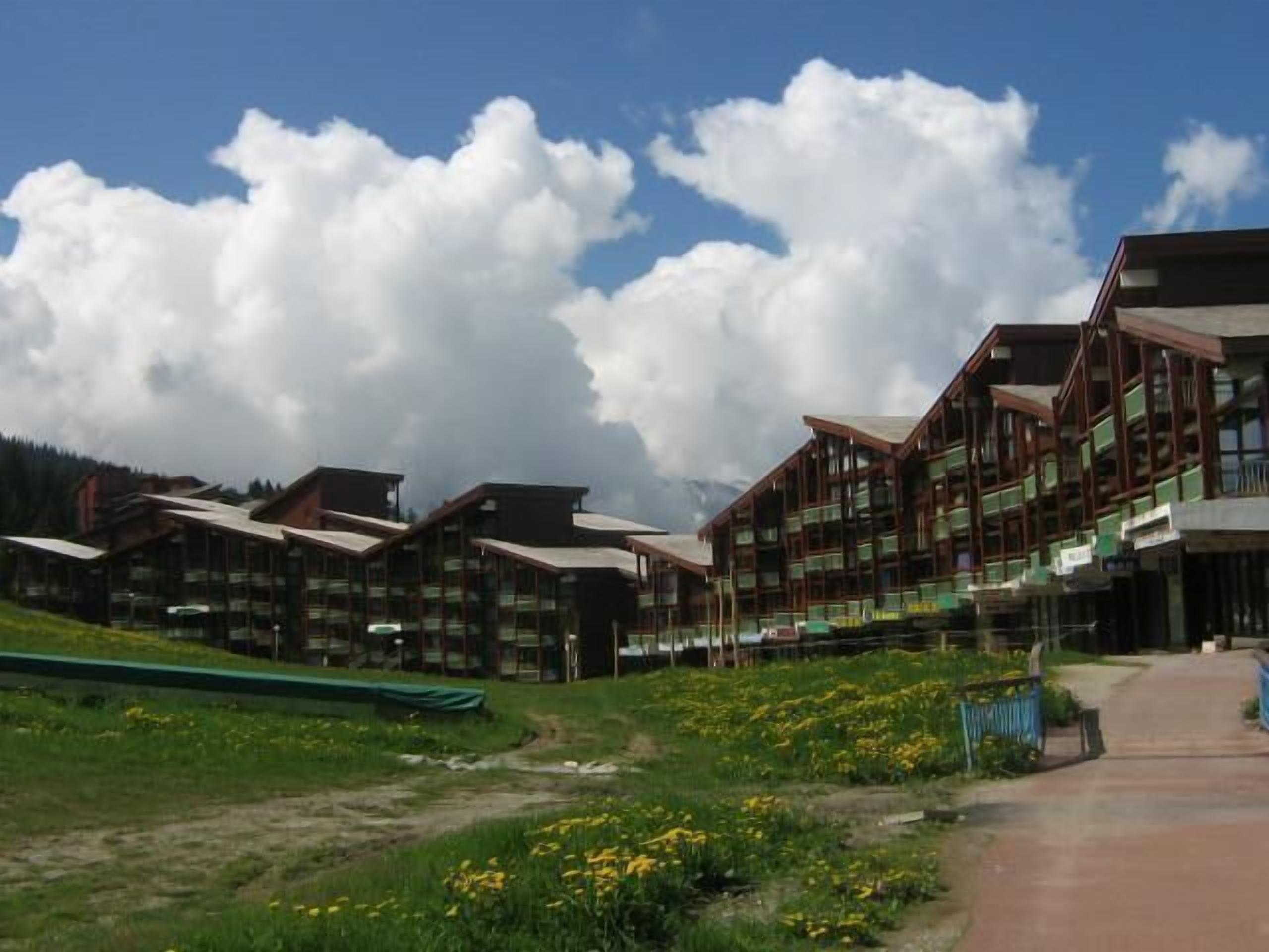 A row of chalet-style buildings with balconies, set against a backdrop of mountains and a clear sky.